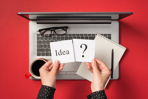 第一人称俯视图的照片，手握两个贴纸的想法和审问标记在开放的灰色笔记本电脑眼镜和红色杯子的饮料孤立的红色背景图片下载