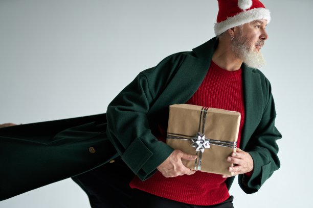 时尚的中年男子戴着圣诞帽，穿着休闲装，看着远处，拿着礼盒，在浅灰色的背景下摆出孤立的姿势图片下载