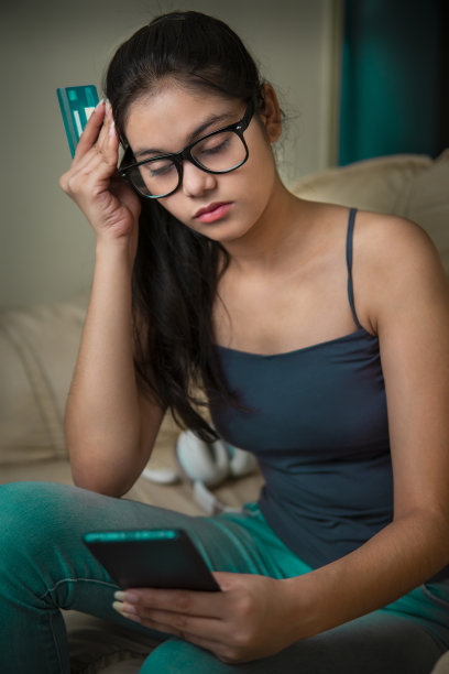 室内形象紧张的亚洲，印度美丽的年轻女子坐在沙发上在客厅的晚上。她在用智能手机进行网上购物时，看到自己的信用卡余额，感到非常震惊。图片下载