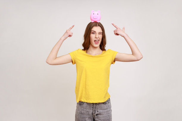 漂亮俏皮的年轻女子，有着愉快的外表和棕色的头发，手里拿着小猪存钱罐，指着它，很高兴有存款。图片下载