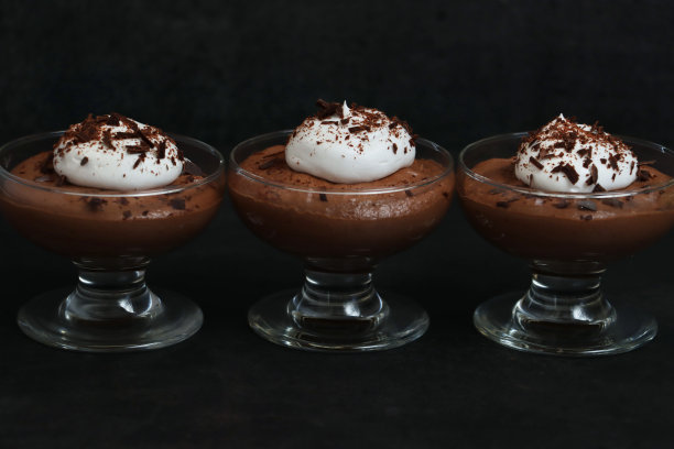 图片是一排三杯自制的巧克力慕斯甜点，上面点缀着生奶油和磨碎的巧克力，背景为黑色，聚焦在前景上图片下载