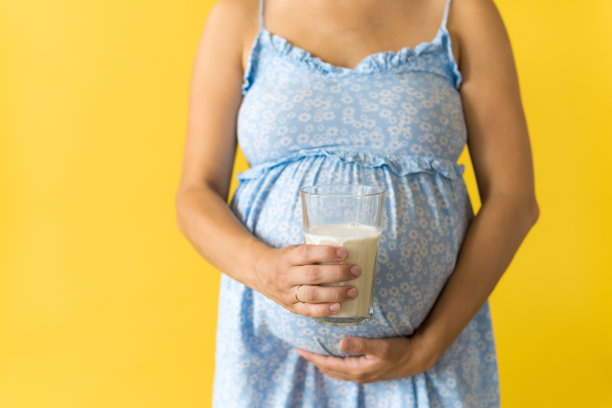 母性，女性气质，维生素，钙，健康的饮食食品，奶制品，炎热的夏天-作物肖像怀孕的不知名的女人在黄色的背景上花蓝色裙子拿着牛奶饮料的杯子图片下载
