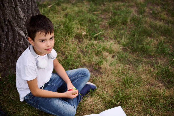 一个英俊的小学生坐在公园的草地上，脖子上戴着耳机，眼睛望向别处，手里拿着一个苹果。小学生放学后休息的照片图片下载