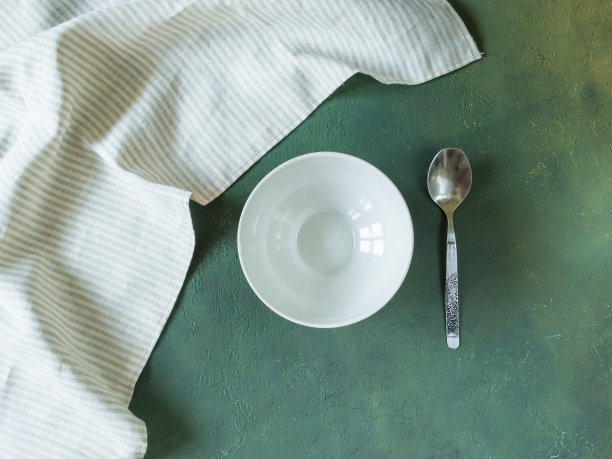 在绿色背景上，有一个空的圆碗在照片的中心。旁边有一把勺子和一张餐巾。你可以把任何菜放在盘子里。水平。俯视图，平躺图片下载