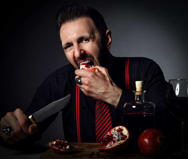 穿着黑色衬衫和吊带的成年男子在桌上端着一瓶和一杯新鲜果汁，咬着石榴图片下载