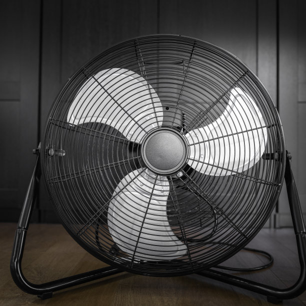 黑色电风扇，有三个灰色的金属叶片。在黑色衣柜前的木地板上用过未插电的电扇。图片下载