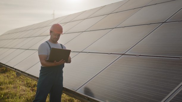 照片中的快乐男工程师戴着防护头盔，手里拿着笔记本电脑，看着相机。身穿制服的英俊男子微笑着站在太阳能发电厂。绿色能源的概念图片下载