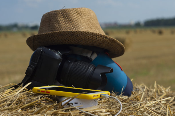 “移动电源”以相机为背景，以干草为背景，以包和帽子为背景，为智能手机充电。便携式旅行充电器。图片下载