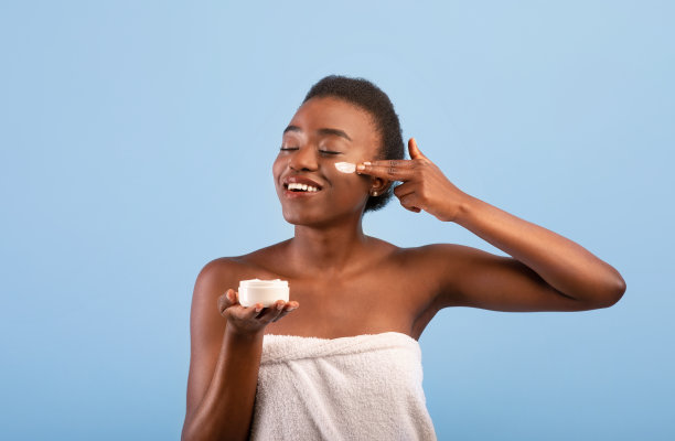 美体的概念。漂亮的非洲裔美国女士在脸上涂奶油，站在蓝色背景下图片下载