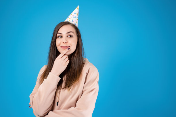 一个年轻漂亮的姑娘戴着节日帽，看上去若有所思。许个生日愿望。孤立在蓝色背景上。副本的空间。图片下载