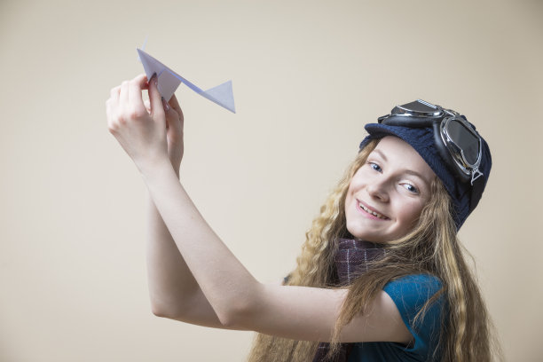 带着飞行眼镜的微笑高加索少女在米色背景上发射折纸飞机。水平射图片下载