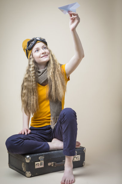 白人少女在飞行眼镜与折纸飞机，而坐在旧的旅行手提箱在米色背景。垂直的图像组成图片下载