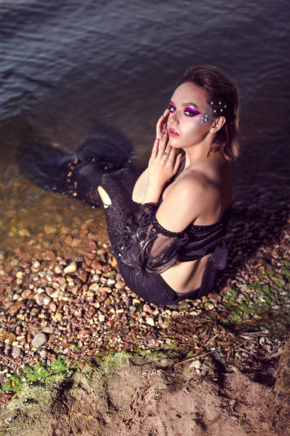 美人鱼在海岸边的岩石上摆姿势，戴着艺术妆容和面部表情，黑色闪亮的尾巴上覆盖着五颜六色的亮片，摆出海洋女王的姿势。垂直拍摄图片下载