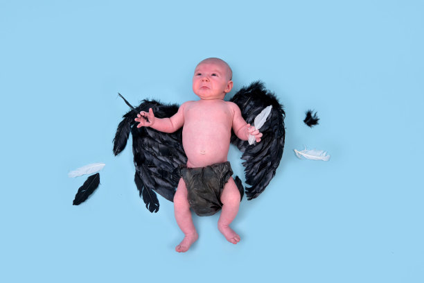 一个带着黑色邪恶翅膀的新生男婴在一个蓝色的工作室背景上，复印空间。一个穿着尿布的白人婴儿一个月大图片下载