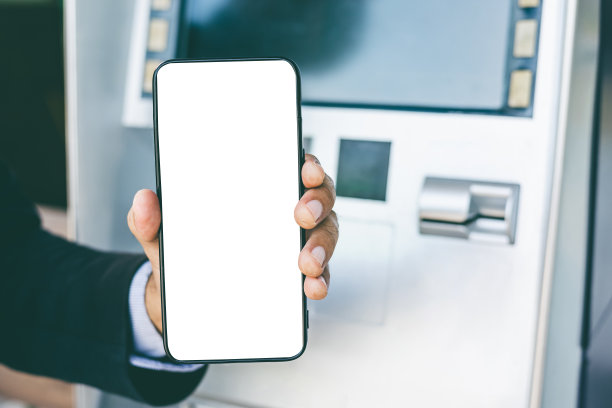 年轻的商人站在自动取款机前用智能手机取钱，而不用自动取款机上的信用卡。他把密码输入智能手机，然后拿走了钱。图片下载