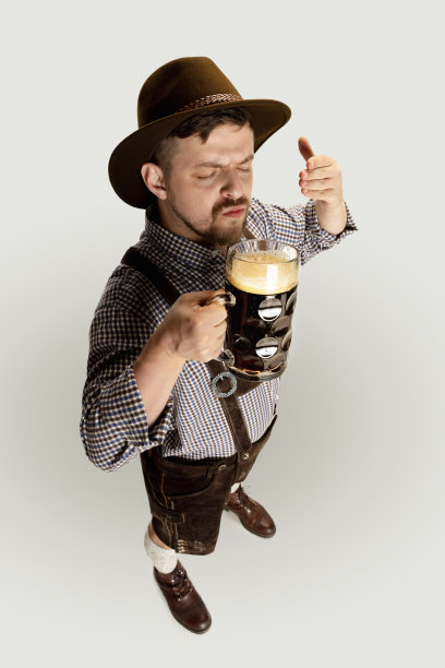 大胡子男子，身着传统奥地利或巴伐利亚服装的侍者，手持马克杯，杯中盛着深黑色的啤酒，背景为灰色图片下载