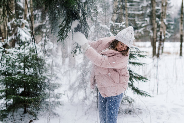 生活方式照片：一位美丽的女孩戴着白色手套、帽子和浅色衣服，在森林里拉着一根被雪覆盖的树枝，雪花落在她身上图片下载