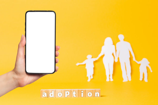在黄色背景上从白纸上剪下的一家人。木立方体与铭文采用。左边，一位女士的手握着一部智能手机。模拟。领养的概念图片下载