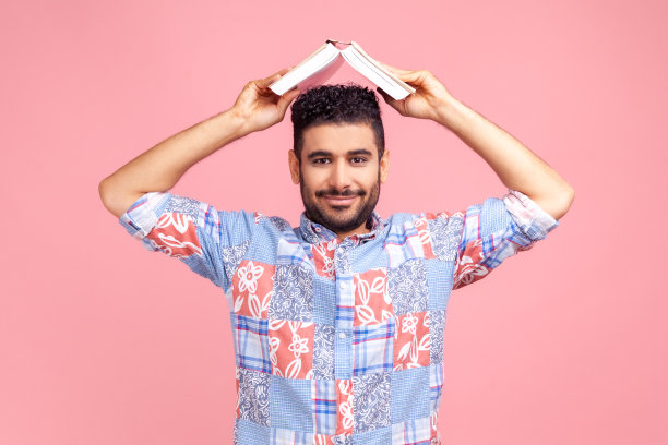 留着胡须，穿着蓝色休闲衬衫，头顶打开的书，面带微笑看着镜头的男子图片下载