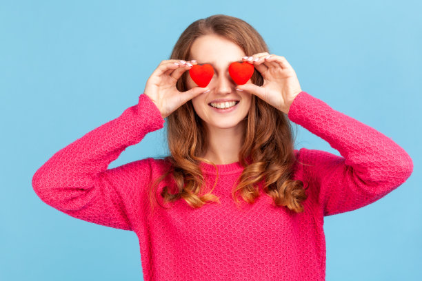 积极的女人，卷发，穿着粉红色的套头衫，对着镜头微笑，用小红心遮住眼睛，表达爱和浪漫的感觉。图片下载