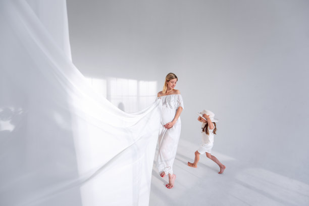 怀孕的母亲持有白色轻质织物在极简室内与光阴影从窗户图片下载