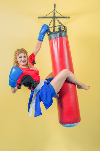 漂亮的年轻女孩穿着武术运动服，拳击手套挂在沙袋上。看镜头。图片下载