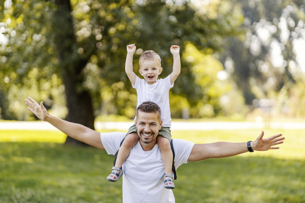 父亲和儿子在大自然中玩耍。当爸爸把男孩扛在肩上时，他们想象着飞机。他们在公园里玩得很开心，穿着同样的休闲服。好家庭的时刻图片下载