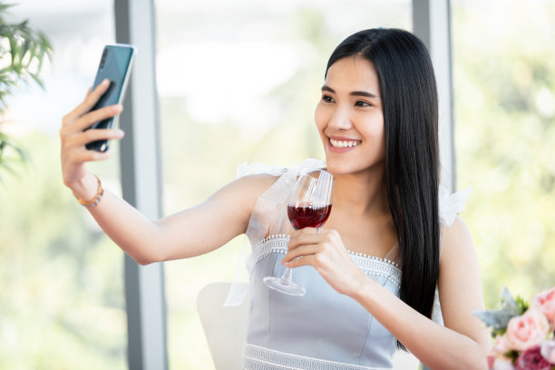 用智能手机自拍的快乐，微笑的亚洲年轻女性坐在餐桌旁，拿着酒杯，餐厅的背景图片下载
