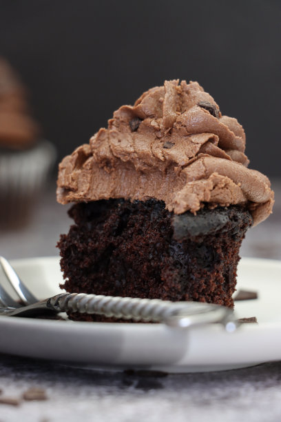 特写图片:白色盘子里装着未包装的纸杯蛋糕，没有咬痕和金属叉子，巧克力黄油糖霜在巧克力块上旋转，黑色背景，聚焦前景图片下载