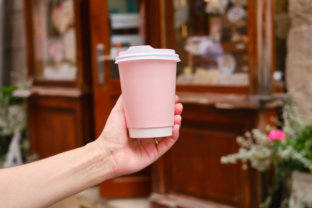 一个女人手里拿着咖啡的粉色纸杯。到城里喝咖啡的时间了。一次性纸杯的城市背景特写。空白的文本，布局。外带咖啡概念图片下载