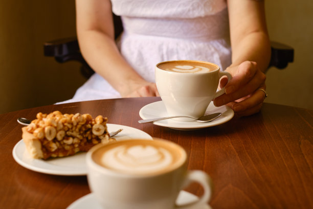 一位女士端着一杯芳香的咖啡和一块甜坚果蛋糕坐在咖啡馆或餐馆里。女孩在早餐后喝一杯热拿铁或卡布奇诺与美味的甜点图片下载