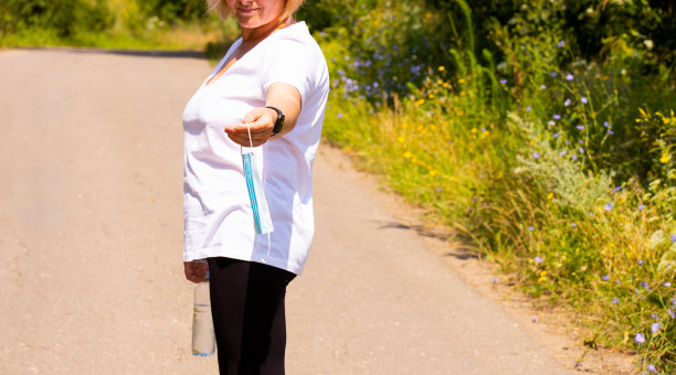 一名健身妇女站在森林的路上，手里拿着一瓶水和医用口罩。图片下载