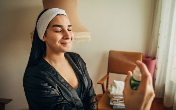 年轻的女人在她的脸上喷洒美容产品在家里库存照片图片下载