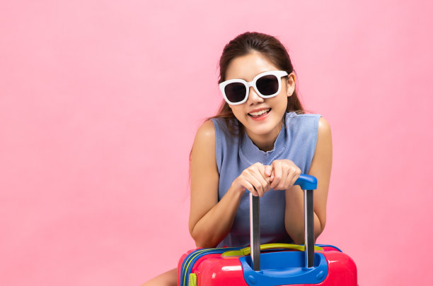 年轻女子提着手提箱，手臂在粉红色的背景下举着。漂亮可爱的梦幻乘客女孩指着copyspace的照片。图片下载