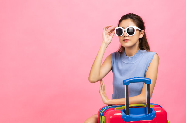 年轻女子提着手提箱，手臂在粉红色的背景下举着。漂亮可爱的梦幻乘客女孩指着copyspace的照片。图片下载