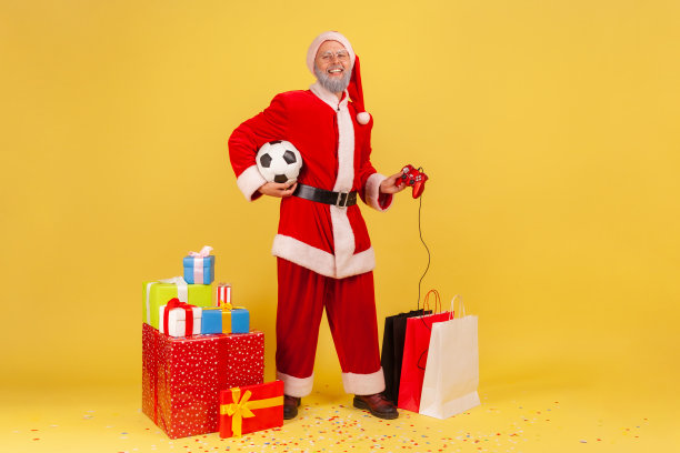 满头灰胡子的老人穿着圣诞老人的服装，脸上露出满意的表情，手里拿着操纵杆和足球，站在礼物旁边。图片下载