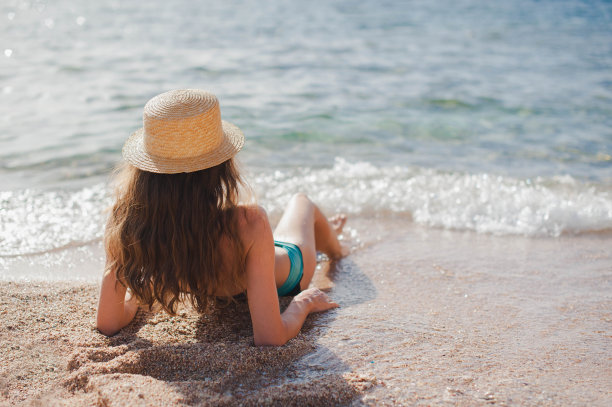 一个戴着帽子的瘦女人躺在海边的黄沙上，靠近海浪。俯视图美丽的女孩在海滩上穿着比基尼。暑期概念旅游图片下载