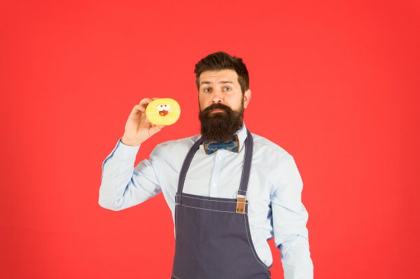 留着胡子的嬉皮士面包师拿着甜甜圈。吃甜甜圈。愉快的心情。甜甜圈的卡路里。釉面甜甜圈。留着胡子穿着围裙卖甜甜圈的男人。甜甜圈的食物。烘焙食品。糖果和蛋糕。垃圾食品图片下载