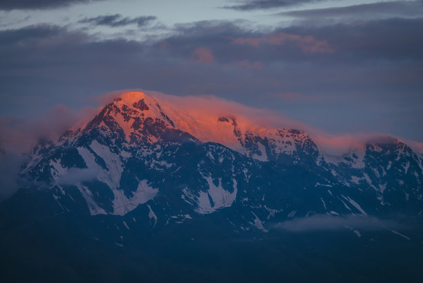 美丽的山景与伟大的雪山被黎明的太阳照亮在低云层。令人敬畏的高山风景与高山的顶峰在日落或日出。上面的大冰川在橙色的灯光下。图片下载
