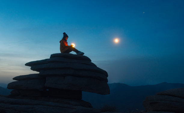 月光下，一个女孩拿着一盏灯，坐在一块石头上图片下载