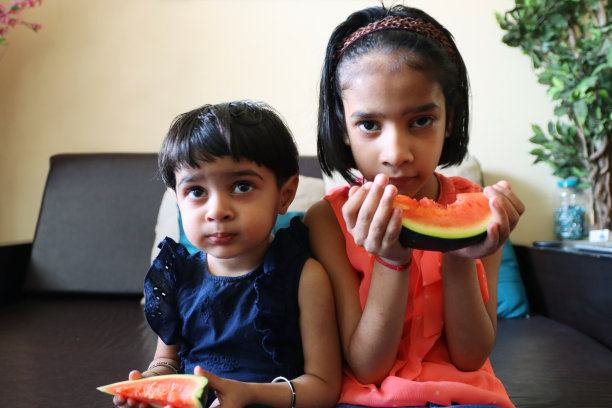 年轻的印度女孩坐在室内的沙发上咬着西瓜片，姐妹们在吃水果图片下载