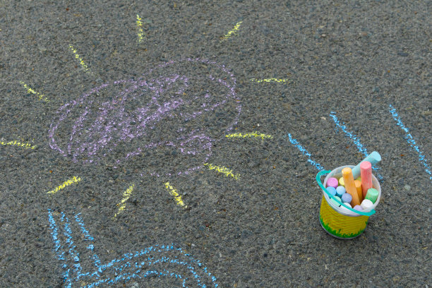 孩子们在柏油路上用粉笔画画。阳光洒在人行道上。画旁边有一个装着蜡笔的桶。图片下载