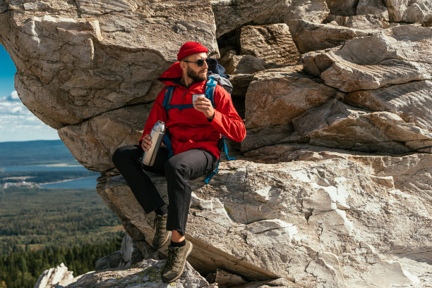 一位男性游客在艰难的爬山之后正在休息。一位游客在山上用保温瓶喝咖啡。一位男性旅行者在山上喝着热茶。本空间图片下载