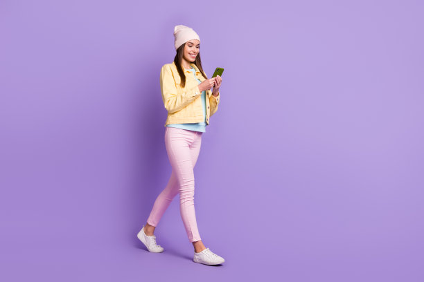 乐观直发女孩的全尺寸侧面照片，去看电话，戴帽子，夹克，裤子，运动鞋孤立在紫色的背景上图片下载