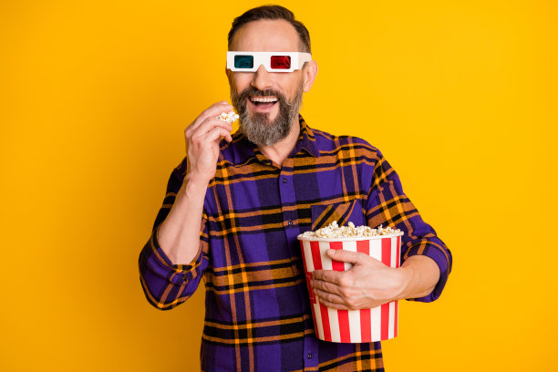欢快快乐的成熟男人的照片，吃爆米花，抱桶，戴眼镜，看电影孤立在黄色背景图片下载