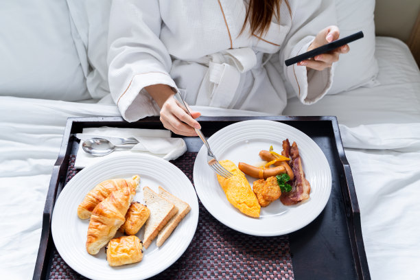美丽的亚洲年轻女子穿着浴袍在卧室的床上吃早餐时使用智能手机图片下载