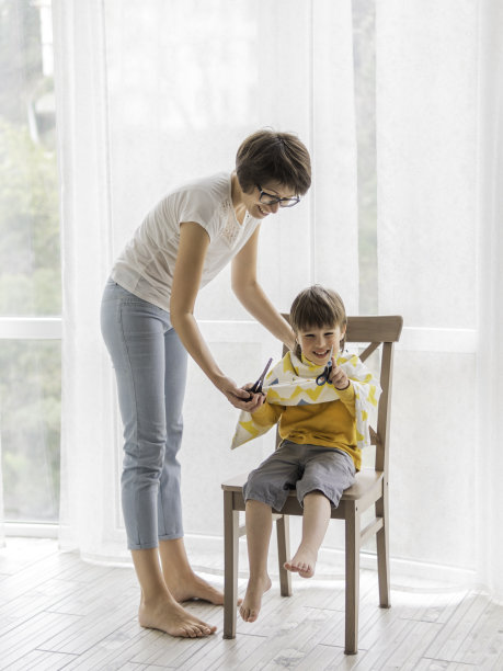 母亲自己给儿子剪头发。小男孩坐着，浑身裹着布，手里拿着剪刀。新冠肺炎疫情隔离和封锁新常态美容家居生活方式。图片下载