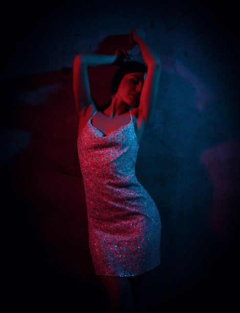 微笑性感美丽的年轻女子在粉红色和蓝色的霓虹灯的背景下，穿着银色的礼服站着和移动图片下载