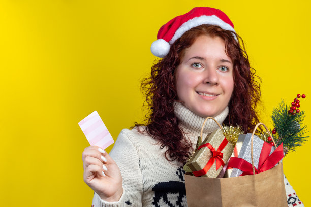 快乐的年轻女人在圣诞帽和斯堪的纳维亚毛衣与信用卡和装满礼物的黄色背景，圣诞节购物，水平，复制空间图片下载