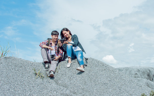 一对亚洲夫妇穿着格子衬衫，背着背包，坐在山顶上，在暑假旅行时查看相机里的照片。图片下载